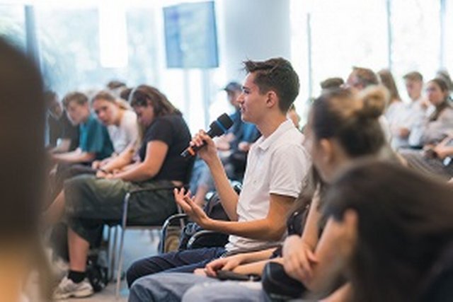 A young man with a microphone speaks while seated among the audience at a conference