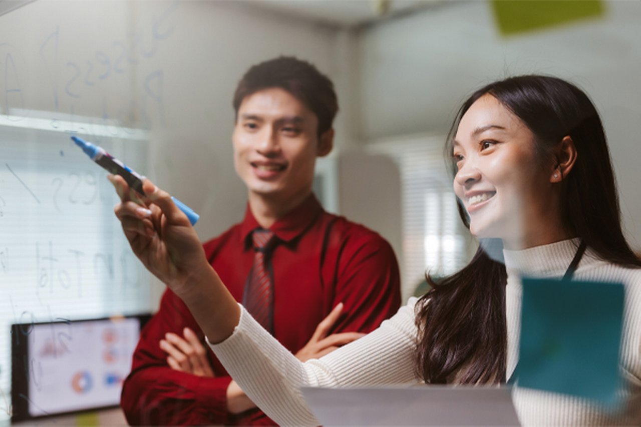 Two people discussing ideas on a clear whiteboard in an office setting.
