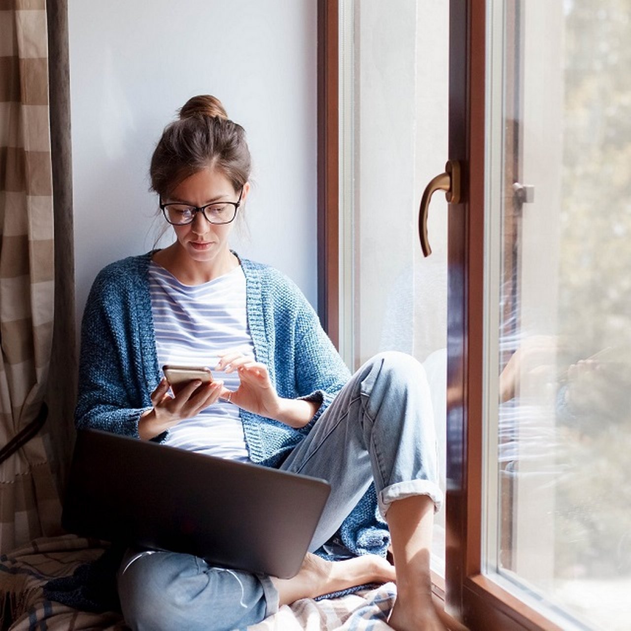 Frau sitzt am Fenster mit Smartphone und Laptop.