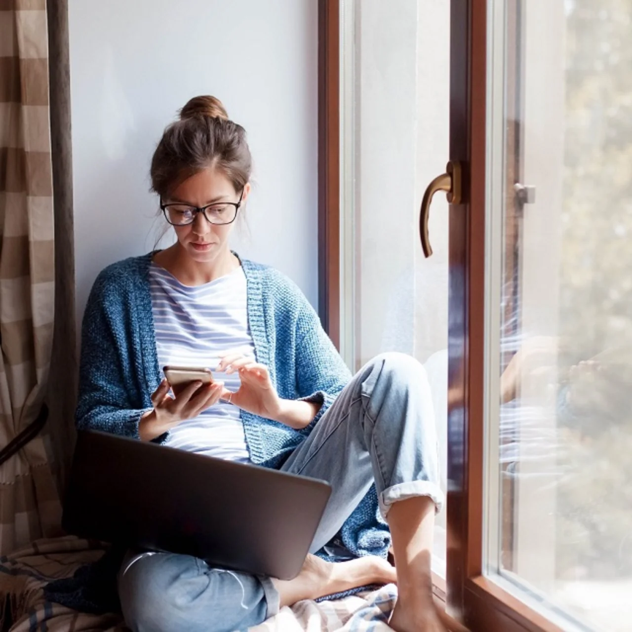 Woman sitting by a window with her mobile phone and a laptop.