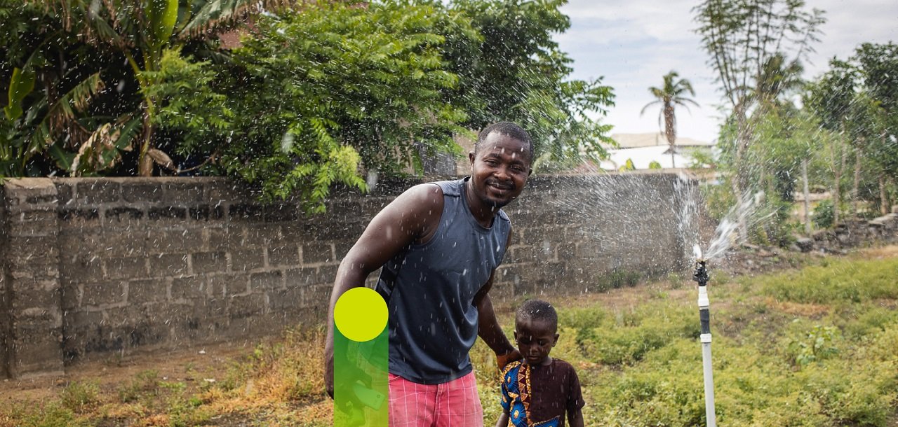 A man and a child are standing on a lawn under a water sprinkler.